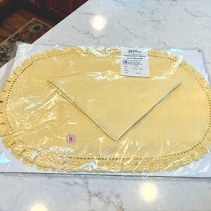 May include: A yellow oval placemat with a decorative scalloped lace border and a matching folded napkin, both enclosed in clear plastic packaging. A white product label is attached to the top, featuring the text Matouk Permanent Press Soil Release.