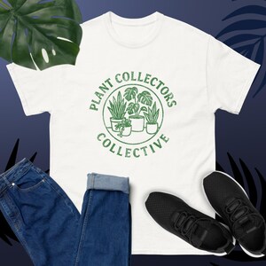 May include: A white t-shirt with a green circular graphic that reads "PLANT COLLECTORS COLLECTIVE" surrounding illustrations of potted plants. The shirt is paired with blue jeans and black sneakers. A large green leaf is in the background.