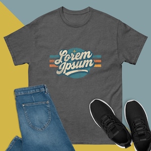Puede incluir: Camiseta gris oscuro con el texto "Lorem Ipsum" en un diseño vintage. La camiseta se muestra con vaqueros azules y zapatillas negras. El texto tiene un estilo retro con acentos azules, naranjas y blancos.