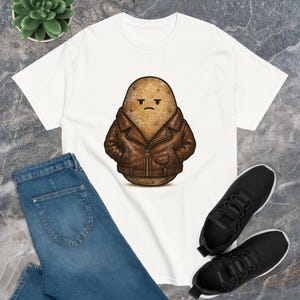 May include: White t-shirt featuring a cartoon potato wearing a brown leather jacket. The potato has a grumpy expression. The shirt is paired with blue jeans and black sneakers.