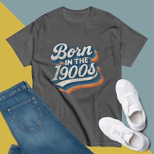 May include: A dark gray t-shirt with the text "Born in the 1900s" in a retro design. The shirt is paired with blue jeans and white sneakers. The text has a vintage look with blue, orange, and white colors.