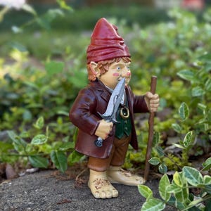 Op de afbeelding: Een grillige tuinkabouterfiguur, met een rode hoed, bruine jas en groen vest. De kabouter houdt een zilveren zwaard en een houten staf vast, staand op een steen. De figuur heeft gedetailleerde gelaatstrekken en is omgeven door groen.