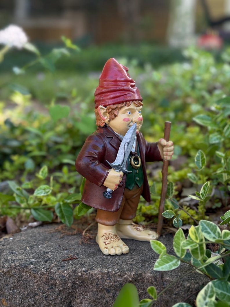Op de afbeelding: Een grillige tuinkabouterfiguur, met een rode hoed, bruine jas en groen vest. De kabouter houdt een zilveren zwaard en een houten staf vast, staand op een steen. De figuur heeft gedetailleerde gelaatstrekken en is omgeven door groen.
