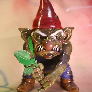 May include: A ceramic garden gnome figurine with an angry expression. The gnome has a red pointed hat, green skin, and is holding a green leaf. It wears blue trousers and brown boots.