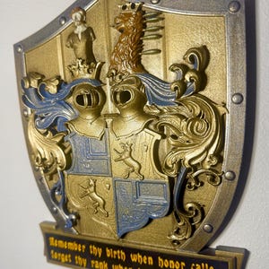 Könnte beinhalten: Ein dekoratives Schild in Gold- und Silberfarben, das ein Wappen mit zwei Rittern, einem Löwen und einer Figur zeigt. Das Schild hat blaue Akzente und ein Banner mit der Aufschrift: "Remember thy birth when honor calls, forget thy rank when humanity speaks."