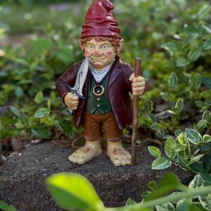 Op de afbeelding: Een grillige tuinkabouterfiguur, met een rode hoed, bruine jas en groen vest. De kabouter heeft een mes in zijn mond en een houten staf. Het beeldje staat op een rots, omringd door groen.