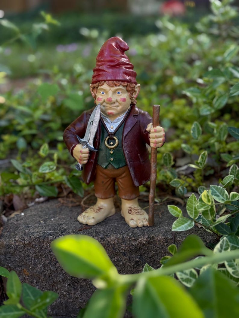 Op de afbeelding: Een grillige tuinkabouterfiguur, met een rode hoed, bruine jas en groen vest. De kabouter heeft een mes in zijn mond en een houten staf. Het beeldje staat op een rots, omringd door groen.