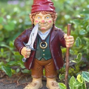 Op de afbeelding: Een grillige tuinkabouterfiguur, met een rode hoed, bruine jas en groen vest. De kabouter houdt een zilveren dolk en een houten staf vast. Een unieke buitendecoratie.