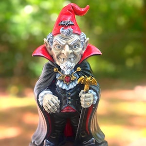 May include: A vampire gnome figurine with a red hat and cape, holding a golden bat-shaped cane. The gnome has a stern expression, a white beard, and is wearing a black suit with a red vest. The base features a bat and skull.
