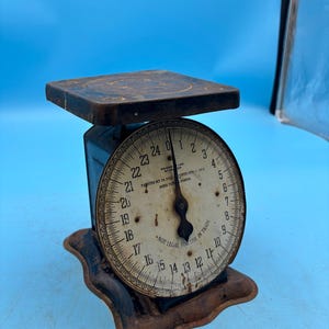 May include: An antique black and brown scale with a circular dial. The dial features numbers and the text "NOT LEGAL FOR USE IN TRADE". The scale has a rectangular platform and a decorative base. The background is a solid blue.