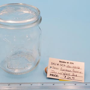 May include: A clear glass jar with a matching lid sits on a light blue surface. A small white tag with handwritten text is next to the jar. A ruler is at the bottom of the image. The jar is labeled "German Garrix Jar w/glass lid".