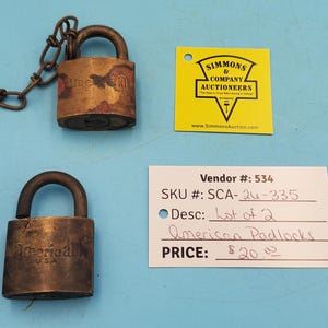 May include: Two antique American padlocks, one with a chain, displayed on a blue surface. The padlocks are made of aged metal, showing signs of wear. A yellow tag and a white label with text are also present.