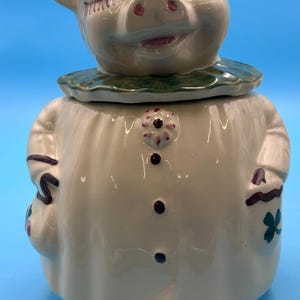 May include: A ceramic cookie jar shaped like a pig wearing a hat and a dress. The pig has closed eyes, a green collar, and a cream-colored body with three dark buttons and a floral accent. A white tag with the number 28 is attached to the base.