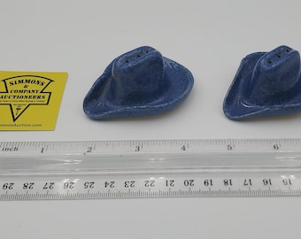 May include: Two blue ceramic cowboy hat-shaped salt and pepper shakers with three holes on top. A yellow Simmons & Company Auctioneers tag and a clear plastic ruler showing measurements in inches and centimeters are placed nearby.