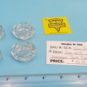 May include: Four clear, hexagonal salt cellars with starburst designs. The salt cellars are on a light blue surface. A yellow tag and a white tag with text are also present.