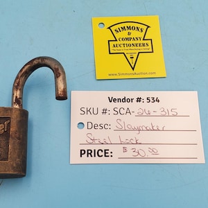 May include: An open, antique steel Slaymaker lock with a weathered appearance. The lock is on a blue surface, with a yellow tag and a white tag with handwritten text. The lock's shackle is raised.
