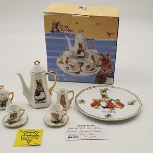 May include: A Peter Rabbit porcelain mini tea set featuring a teapot, sugar bowl, creamer, two cups with saucers, and a plate. Each piece is decorated with illustrations of Peter Rabbit and gold trim. The set is displayed with its original box.