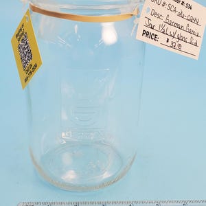 May include: A clear glass jar with a glass lid. The jar has a gold-colored band around the top. A tag attached to the jar reads "Jar 1/2 gal w/ glass lid". A ruler is at the bottom of the image.