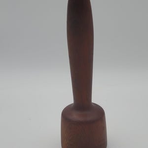 May include: A dark brown wooden mallet with a long, tapered handle and a cylindrical head. A small black metal hook is attached to the top of the handle. The mallet is positioned vertically, showcasing its smooth, polished wood grain texture.