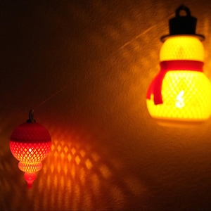 May include: A string of decorative lights, including a snowman-shaped lantern with a red scarf and top hat. Other lanterns are red and orange, casting warm light and shadows on the wall. The lights are ideal for festive decor.
