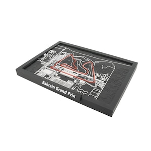 May include: A black framed relief map of the Bahrain Grand Prix race track. The track layout is highlighted in red, with white detailing of the surrounding infrastructure. The text "Bahrain Grand Prix" is visible on the frame.