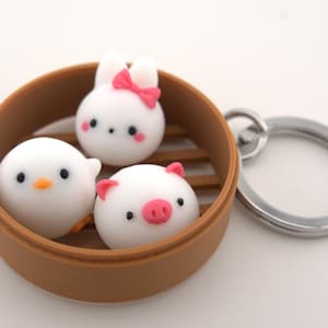 May include: A novelty keychain featuring three white, round, cartoon characters inside a brown bamboo steamer. The characters include a chick with an orange beak, a bunny with a pink bow, and a pig with a pink snout. A silver keyring is attached.