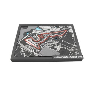 May include: A detailed, three-dimensional map of the United States Grand Prix race track, framed in a dark gray border. The track is highlighted in red, with surrounding infrastructure in white and gray. The text "United States Grand Prix" is visible.