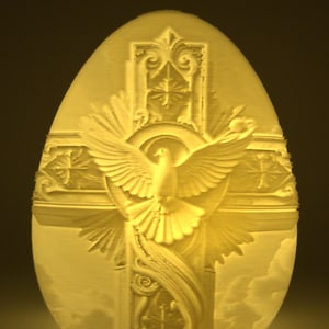May include: A cream-colored, egg-shaped sculpture featuring a dove with outstretched wings, set against a cross and a backdrop of clouds. The design includes ornate details and a halo around the dove's head.