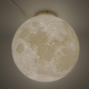 May include: A spherical, off-white ceiling light fixture resembling the moon. The light has a textured surface with craters and a soft glow, creating a celestial ambiance. The fixture is suspended from the ceiling with a thin white cord.