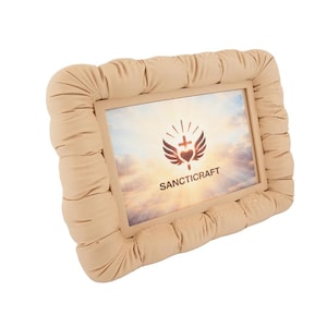 May include: A beige, rectangular picture frame with a padded, quilted border. The frame holds an image with a logo that includes a heart, cross, and wings, with the word "SANCTICRAFT" below. The background of the image is a cloudy sky.