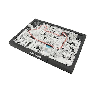 May include: A 3D model of Las Vegas, Nevada, in a black frame. The map features white buildings and streets, with a red outline highlighting a specific route. The words "Las Vegas" are printed on the frame.