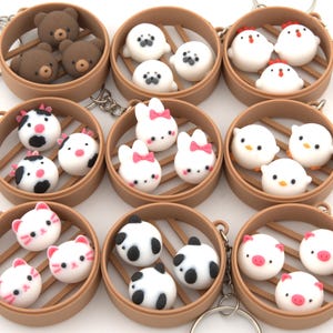 May include: A collection of novelty keychains featuring miniature animal-shaped dumplings in bamboo steamer baskets. The keychains include bear, seal, chicken, cow, bunny, cat, panda, and pig designs, each with detailed facial features and colorful accents.