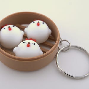 May include: A novelty keychain featuring three white, chicken-shaped dumplings inside a brown bamboo steamer. Each dumpling has a red comb and beak. The keychain includes a silver-colored metal ring.