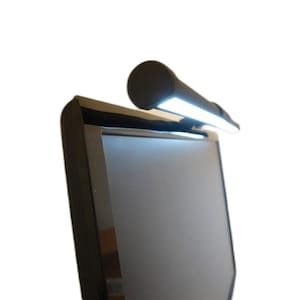 May include: A black LED monitor light bar is mounted on top of a black computer monitor. The light bar is cylindrical and emits bright white light. The monitor is rectangular with a black frame.