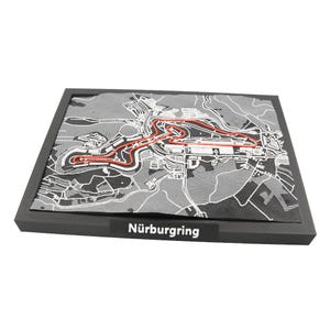 May include: A 3D map of the Nürburgring race track, featuring a red track outline against a gray and white topographical background. The map is set in a black frame, with the track name printed on the front.