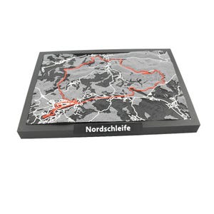 May include: A gray and black relief map of the Nordschleife race track, with a red track outline and white road details. The map is set in a dark gray frame, with the word "Nordschleife" printed on a black label.