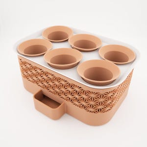 May include: A terracotta-colored planter with six circular pot holders. The planter has a geometric pattern on the sides and a small rectangular compartment. The pot holders are arranged on a white tray.