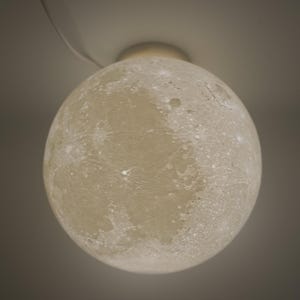 May include: A spherical, off-white ceiling light fixture resembling the moon. The light has a textured surface with craters and other lunar details. The fixture is attached to the ceiling with a white cord.
