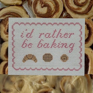 May include: A rectangular needlepoint canvas featuring the pink stitched text "i'd rather be baking" surrounded by a pink scalloped border, with small brown stitched icons of a croissant, a cookie, and a muffin positioned below the text.