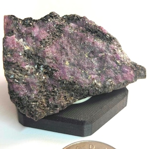 Puede incluir: Una gema en bruto con una mezcla de tonos gris oscuro y rosa vibrante. La piedra descansa sobre un pequeño soporte rectangular negro. Una moneda canadiense está en primer plano.