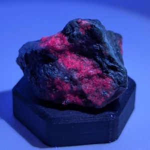Puede incluir: Una roca oscura y rugosa con una vibrante coloración roja y negra, exhibida sobre un soporte hexagonal negro. La superficie de la roca tiene una apariencia texturizada, con las áreas rojas que parecen brillar. El fondo es azul sólido.
