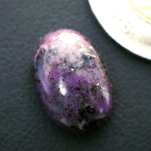 Puede incluir: Una gema ovalada pulida, de color púrpura vibrante, salpicada de motas negras y doradas. La piedra se encuentra sobre un fondo oscuro, con una porción de una moneda de plata visible en la esquina superior derecha.