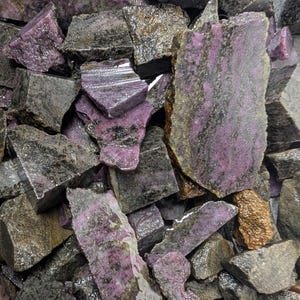 Peut inclure: Un ensemble de pierres brutes avec un mélange de couleurs. Les pierres ont un extérieur gris foncé et brun avec des taches de violet vif. La texture semble rugueuse et naturelle, avec des structures cristallines visibles.