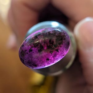 Puede incluir: Primer plano de una piedra preciosa pulida de forma ovalada. La piedra presenta un patrón arremolinado de tonos morados oscuros y negros. La piedra preciosa está engastada en un engaste de metal plateado, que refleja la luz.