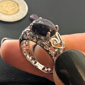 May include: A silver ring featuring a dark purple oval gemstone. The ring's band has an intricate, filigree pattern. A coin is visible in the background.