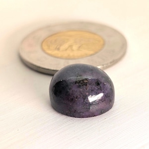 Puede incluir: Una gema pulida de color púrpura oscuro con forma de cúpula redondeada. La piedra tiene una superficie lisa y reflectante. Una moneda de plata con un centro dorado está en el fondo, sobre un fondo blanco.