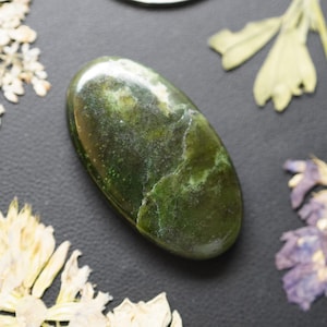 May include: An oval-shaped polished gemstone in a deep forest green colour with lighter green and white mineral inclusions throughout its smooth, glossy surface, showcasing natural variations in the stone's texture and colour depth.