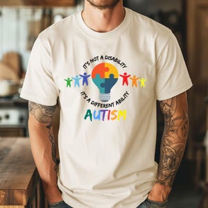 May include: Cream-colored t-shirt with a colorful graphic promoting autism awareness. The design includes a lightbulb shape with a puzzle piece, stick figures, and the text "It's not a disability, it's a different ability. Autism."