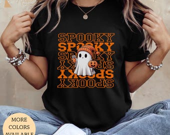 May include: Black t-shirt with the word "SPOOKY" repeated in orange, featuring a cartoon ghost wearing an orange beanie and holding a pumpkin. The shirt also has the text "MORE COLORS AVAILABLE".