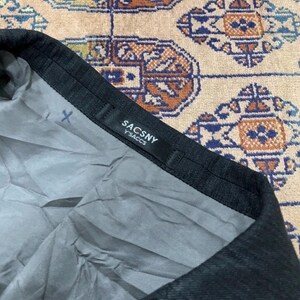 May include: Close-up of a dark gray garment with a black label that reads "SACSNY Y'SACCS". The interior lining is a light gray color. The garment is on a patterned beige and brown surface.